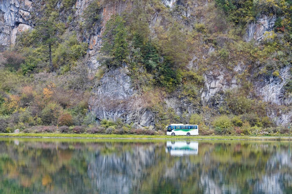 Bus traveling along a tranquil lake reflecting mountain cliffs and lush greenery.