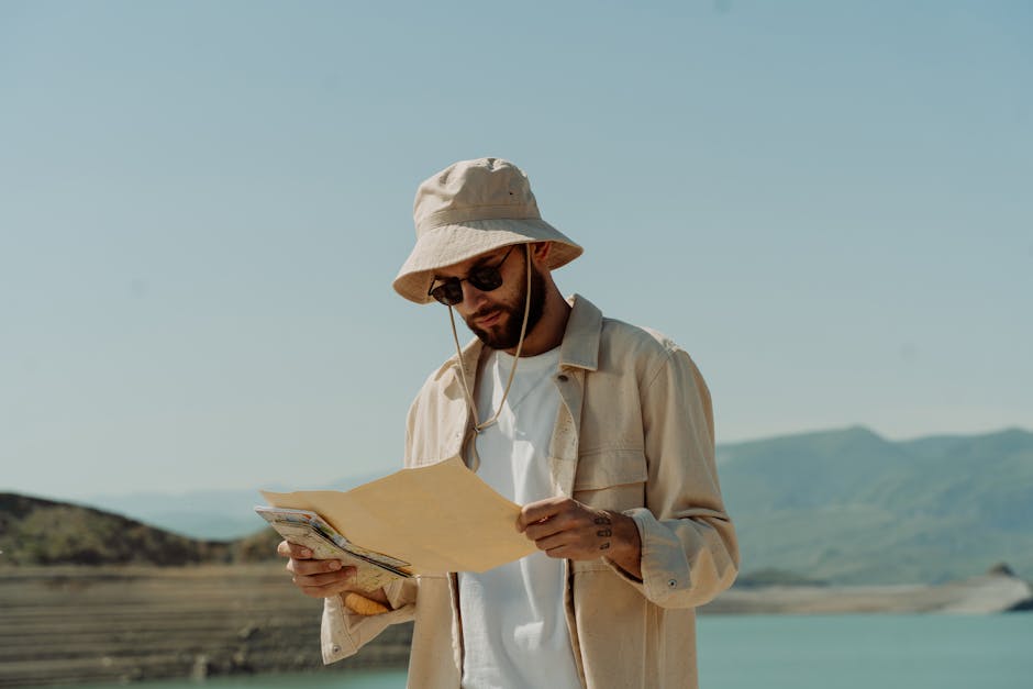 Man in a bucket hat and sunglasses studies a map outdoors by a scenic lake.